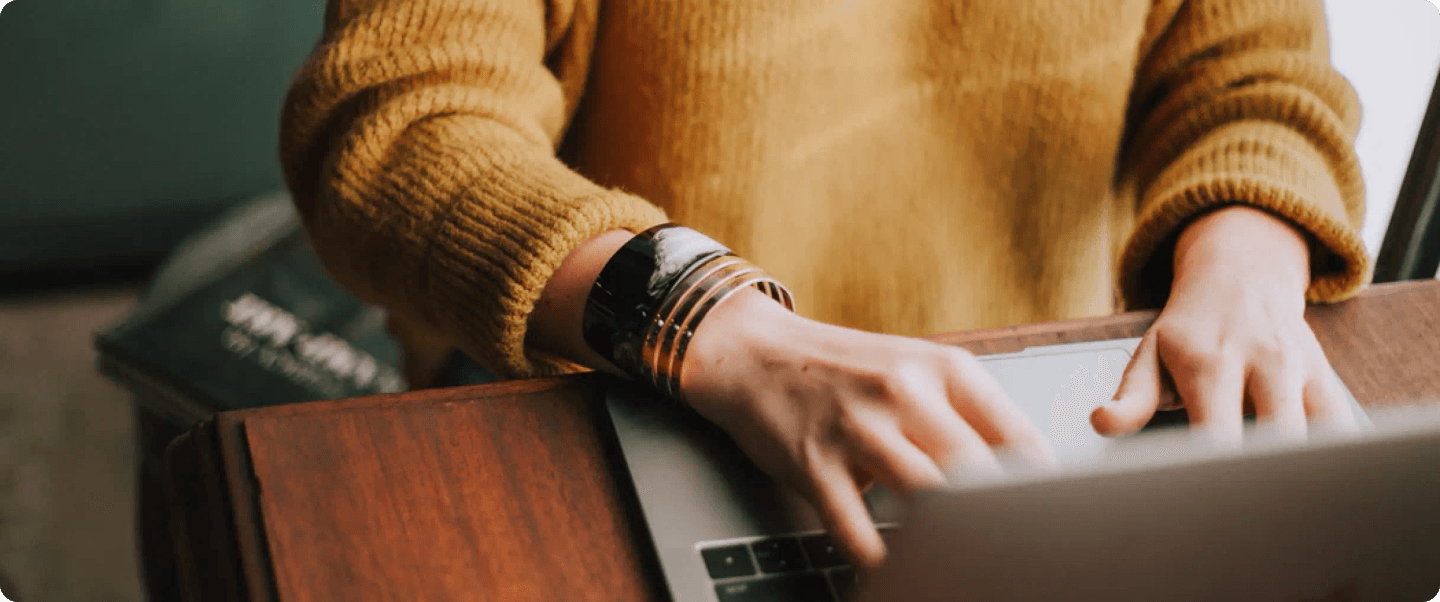 A person in a yellow sweater typing on a laptop at a wooden desk.