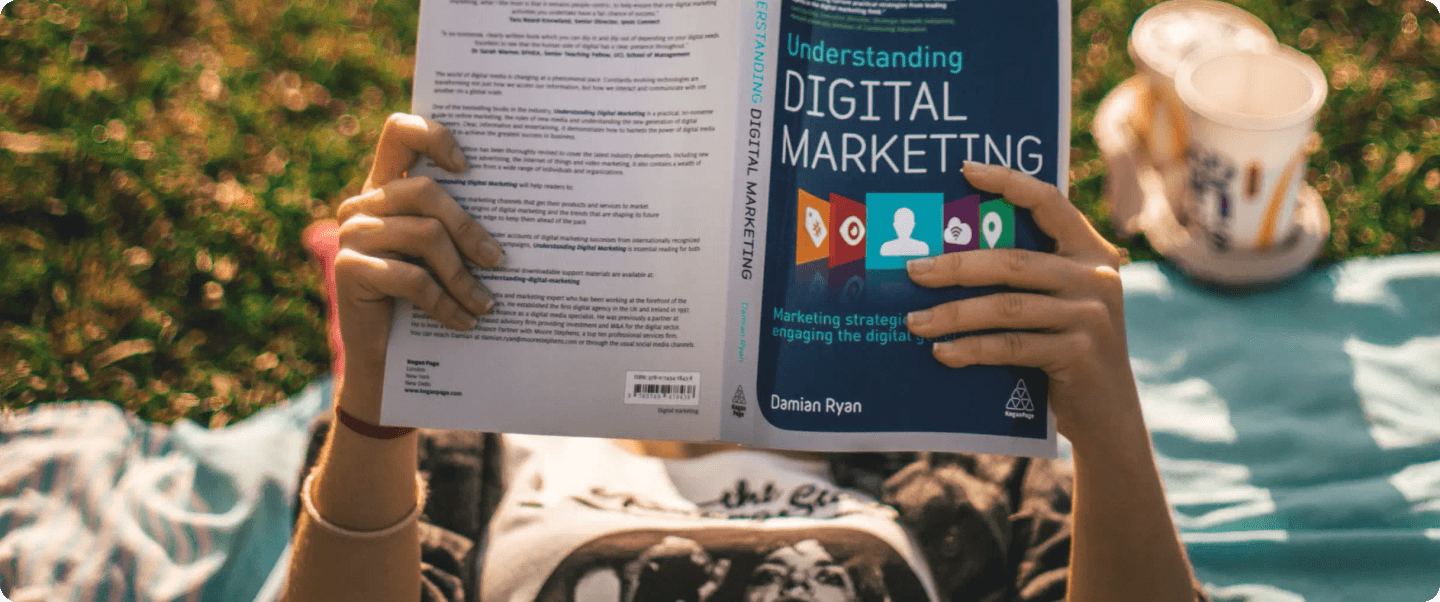 A person lying on a picnic blanket, reading a book about digital marketing.